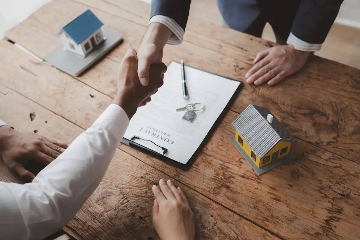 Deux personnes qui se serrent la main au dessus d'un contrat et d'une maquette de maison posée sur la table